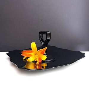 May include: A black, square serving tray with a scalloped edge and a black handle. A single orange and yellow flower is placed on the tray.