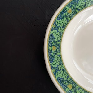 May include: A white ceramic plate with a green and blue floral pattern around the edge. The plate has a raised rim and a shallow well in the center.