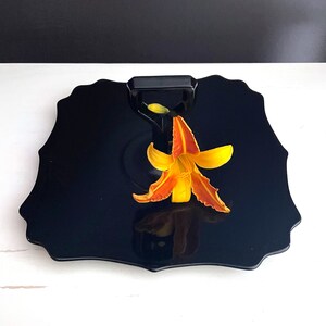 May include: A black, scalloped serving tray with a black handle and a single orange lily flower on top.