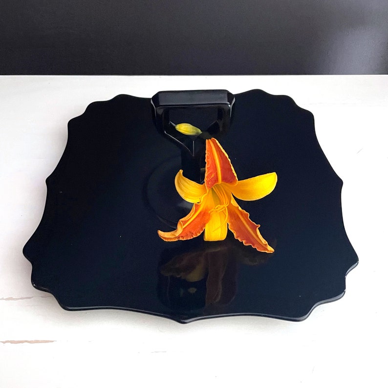 May include: A black, scalloped serving tray with a black handle and a single orange lily flower on top.