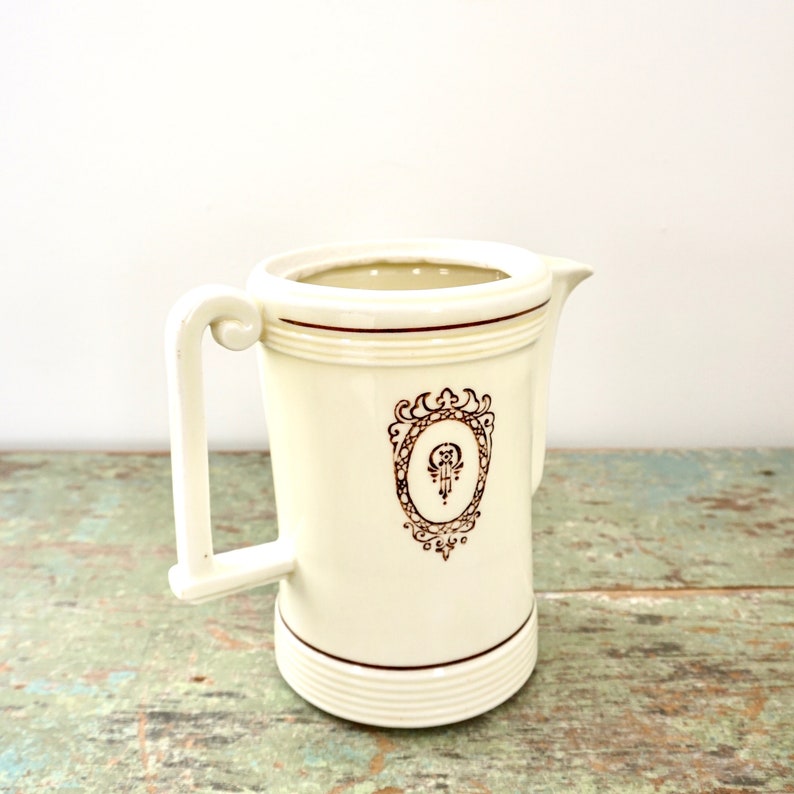 May include: A white ceramic creamer with a brown ornate design. The creamer has a handle and a spout.