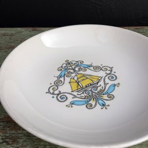May include: White ceramic plate with a decorative nautical design. The design features a yellow sailboat, blue floral accents, and swirling gray lines. The plate has a slightly curved rim.