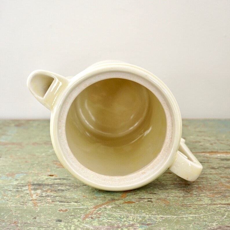 May include: A vintage cream-colored ceramic pitcher with a spout and a handle. The pitcher is empty and has a smooth, glazed finish.