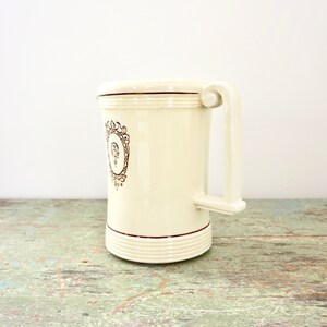 May include: A white ceramic mug with a brown handle and a gold design. The design features a crest with a letter "Q" inside.