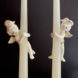 Peut inclure: Une paire de bougeoirs en céramique blanche avec des figurines d'anges tenant les bougies. Les anges ont des accents dorés et sont en position de sommeil.