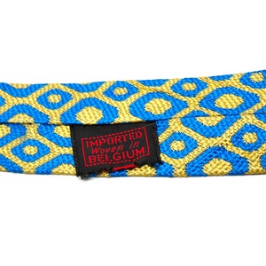 Puede incluir: Primer plano de una corbata estampada con un dise&ntilde;o geom&eacute;trico azul y dorado repetido. La corbata tiene una etiqueta que dice "Importado tejido en B&eacute;lgica". El fondo es blanco.