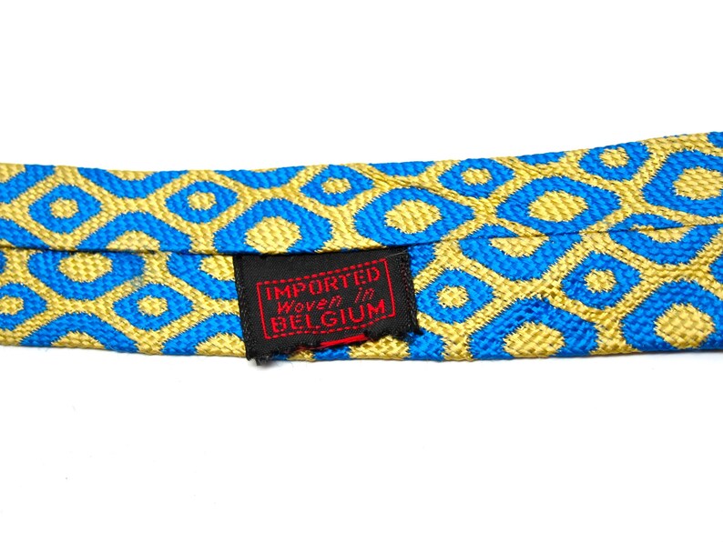 Puede incluir: Primer plano de una corbata estampada con un dise&ntilde;o geom&eacute;trico azul y dorado repetido. La corbata tiene una etiqueta que dice "Importado tejido en B&eacute;lgica". El fondo es blanco.