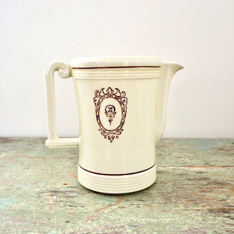 May include: A white ceramic pitcher with a brown decorative design. The pitcher has a handle and a spout. The design features a stylized floral motif with a central oval.