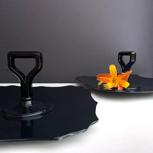 May include: Two black glass serving trays with a scalloped edge. One tray has a black handle in the shape of a spade. The other tray has a single orange flower on top.