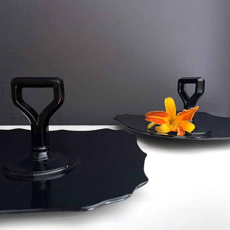 May include: Two black glass serving trays with a scalloped edge. One tray has a black handle in the shape of a spade. The other tray has a single orange flower on top.
