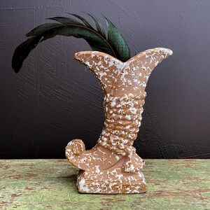 May include: A brown ceramic vase with white and gold speckles. The vase is shaped like a horn and has a decorative base. A green feather is placed inside the vase.