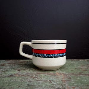 May include: A white ceramic coffee mug with a red and blue geometric design. The mug has a rounded shape and a handle.