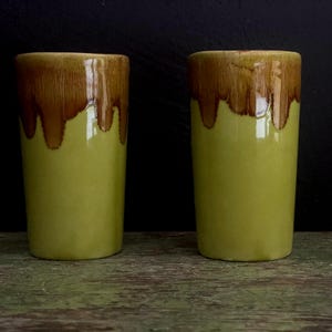 May include: Two green ceramic cups with brown glaze dripping down the sides. The cups are sitting on a wooden surface.