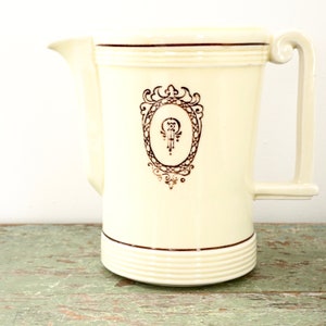 May include: A white ceramic pitcher with a gold-colored design. The design features a decorative oval frame with a stylized letter "O" inside. The pitcher has a gold-colored rim and a gold-colored handle.