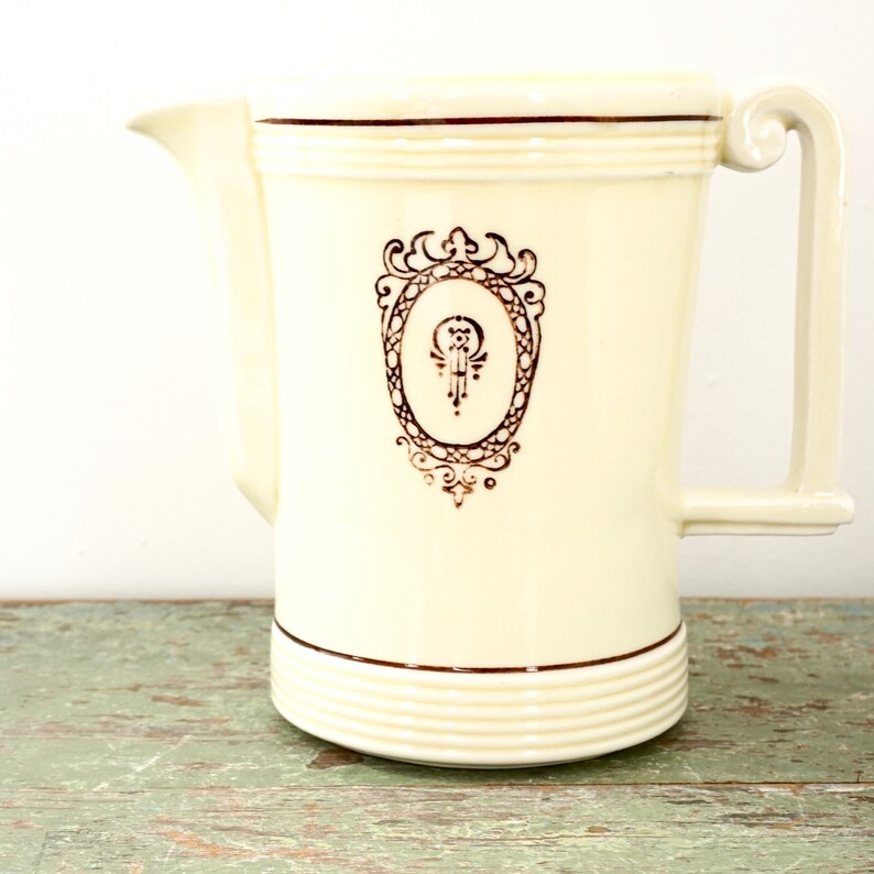May include: A white ceramic pitcher with a gold-colored design. The design features a decorative oval frame with a stylized letter "O" inside. The pitcher has a gold-colored rim and a gold-colored handle.