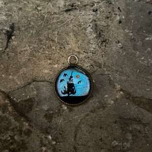 May include: A round pendant featuring a blue butterfly wing design. The pendant depicts a silhouette of a tree and birds against a blue background. The pendant is framed in a silver-toned metal.