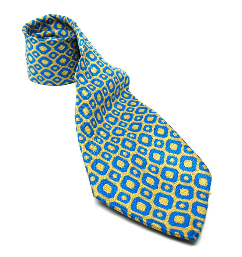 Puede incluir: Una corbata estampada en azul y dorado. La corbata presenta un dise&ntilde;o geom&eacute;trico repetitivo de formas azules sobre un fondo dorado. La corbata est&aacute; enrollada en la parte superior e inclinada hacia abajo, mostrando el patr&oacute;n y la textura.
