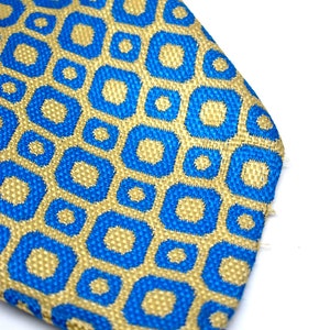 Puede incluir: Primer plano de una corbata estampada. La corbata presenta un dise&ntilde;o geom&eacute;trico repetitivo de cuadrados y c&iacute;rculos azules sobre un fondo dorado. La tela tiene una apariencia texturizada y la corbata est&aacute; sobre un fondo blanco.