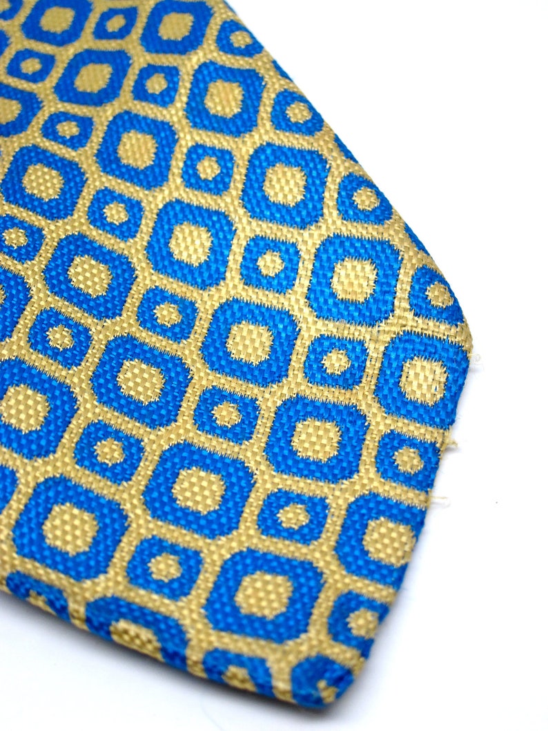 Puede incluir: Primer plano de una corbata estampada. La corbata presenta un dise&ntilde;o geom&eacute;trico repetitivo de cuadrados y c&iacute;rculos azules sobre un fondo dorado. La tela tiene una apariencia texturizada y la corbata est&aacute; sobre un fondo blanco.