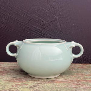 Puede incluir: Una jarra de crema de cerámica azul claro con dos asas. La jarra tiene un cuerpo redondeado y un acabado liso y brillante.