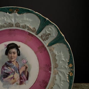 May include: A decorative porcelain plate with a portrait of a woman in a kimono holding a flower. The plate features a pink and white center, a green and gold rim, and ornate detailing.