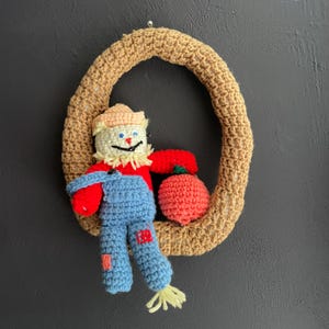 May include: A handmade crochet wreath featuring a scarecrow figure and a red apple. The scarecrow has a beige hat, a straw-like face, a red shirt, and blue overalls. The wreath is made of tan yarn.