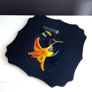 May include: A black, scalloped serving tray with a flower and a black metal handle. The flower is orange and yellow with a green stem.