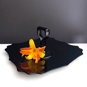 May include: A black serving tray with a scalloped edge and a black handle. A single orange flower is placed on the tray.