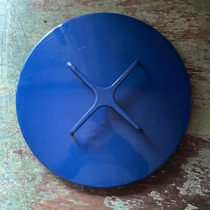 May include: A round, cobalt blue metal lid with a cross-shaped handle. The lid has a glossy finish and appears to be a part of a larger piece of furniture or decor. The handle is attached to the center of the lid.