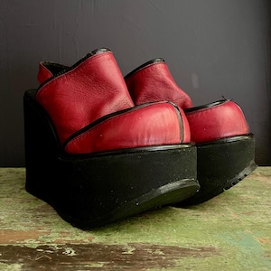 May include: A pair of red leather platform sandals with black soles and a slingback design. The sandals have a thick, black platform sole and a red leather upper with black trim. The shoes are a bold fashion statement.