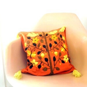 May include: Orange throw pillow with embroidered floral and bird design in yellow, brown, and black. The pillow has yellow tassels on each corner.