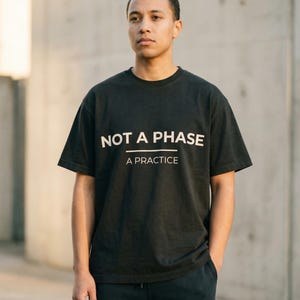 Könnte beinhalten: Schwarzes T-Shirt mit dem weißen Text "NOT A PHASE" über "A PRACTICE". Die Person trägt eine schwarze Hose und steht vor einer Betonmauer, die Hände in den Taschen.
