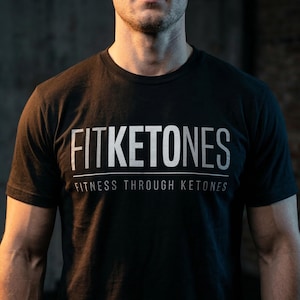 May include: Black crew neck t-shirt with the white text "FITKETONES" above "FITNESS THROUGH KETONES". The shirt is made of a soft material and is suitable for workouts or casual wear.