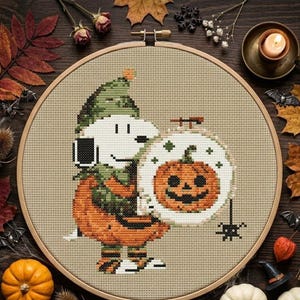 Puede incluir: Un bordado de punto de cruz en un bastidor de madera circular que muestra a un perro blanco con un gorro verde y un disfraz de calabaza, sosteniendo un bastidor más pequeño con una cara de calabaza y una pequeña araña negra colgando.