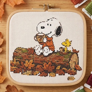May include: A cross-stitch embroidery in a square wooden hoop featuring Snoopy wearing an orange sweater and holding a mug while sitting on a log with Woodstock, surrounded by fallen autumn leaves on a white fabric background.