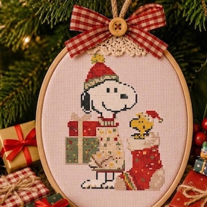 May include: A circular wooden embroidery hoop containing a cross-stitch design of Snoopy wearing a Santa hat and a patterned sweater, holding a gift, next to Woodstock in a red stocking. A red and white gingham bow with a wooden button sits on top.