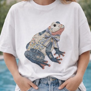 May include: White t-shirt featuring a patchwork frog design. The frog is made from denim, floral, and gingham fabrics, with a blue bow tie. The frog's body is a mix of denim and floral patterns, with red gingham accents on the feet.