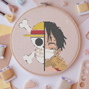 Puede incluir: Un bastidor de bordado circular de madera que contiene un diseño de punto de cruz dividido por la mitad. El lado izquierdo presenta una calavera blanca con huesos cruzados y un sombrero rojo y amarillo, mientras el derecho muestra un personaje.