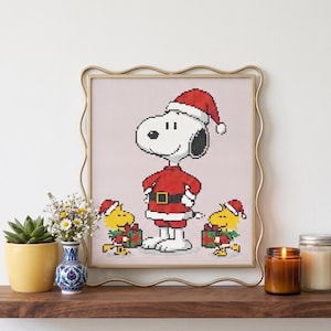 Puede incluir: Un patrón de punto de cruz que muestra a un perro blanco con gorro y traje de Papá Noel, flanqueado por dos pequeños pájaros amarillos que también llevan gorros navideños y sostienen regalos verdes, en un marco dorado ondulado.