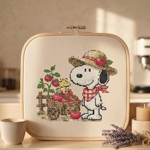 May include: A square wooden embroidery hoop containing a cross-stitch design of Snoopy wearing a straw hat and a red chequered neckerchief, standing next to a wooden cart filled with red tomatoes and a small yellow bird sitting on top.