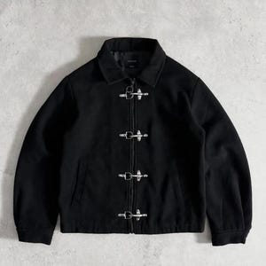 May include: Black jacket with a collared neckline and silver toggle closures. The jacket has a simple design with a clean, modern aesthetic. The jacket is made of a soft, matte material.