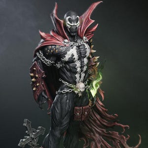 Puede incluir: Figura detallada del personaje de cómic Spawn. La figura es predominantemente negra con un emblema blanco en el pecho, una capa roja y cadenas plateadas. La figura sostiene un efecto de energía verde y se encuentra sobre una base con una cruz.