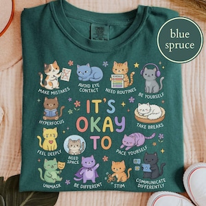 May include: A blue spruce t-shirt featuring a graphic of various cats with the text IT'S OKAY TO and phrases including MAKE MISTAKES, AVOID EYE CONTACT, NEED ROUTINES, BE YOURSELF, HYPERFOCUS, TAKE BREAKS, FEEL DEEPLY, NEED SPACE, PACE YOURSELF, UNMASK, BE DIFFERENT, STIM, and COMMUNICATE DIFFERENTLY.