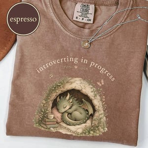 May include: An espresso-colored t-shirt with a crew neckline and a Comfort Colors label. The graphic features a sleeping green dragon inside a cave with books and a mug, accompanied by the text "introverting in progress" in a light font.