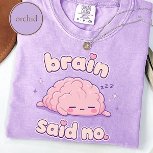 May include: An orchid-colored short-sleeve t-shirt featuring a graphic of a sleeping pink brain with the text "brain Said no." and small "zzz" symbols. The shirt has a crew neckline, a Comfort Colors brand label, and a small silver-toned necklace.