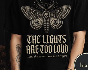 May include: A black t-shirt featuring a graphic of a moth with a skull on its thorax. The text reads THE LIGHTS ARE TOO LOUD (and the sounds are too bright) in a gothic font. The design is centered on the chest in shades of white and gray.