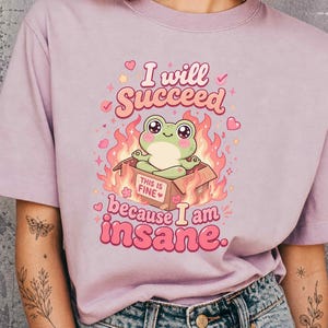 May include: A light purple short-sleeve t-shirt featuring a graphic of a green frog sitting in a cardboard box labeled THIS IS FINE, surrounded by pink flames and hearts, with the text I will SUCCEED because I am insane printed in pink.