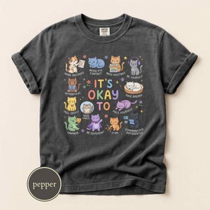 May include: A charcoal gray t-shirt featuring a central graphic of various colorful cartoon cats with the text IT'S OKAY TO, accompanied by phrases like MAKE MISTAKES, AVOID EYE CONTACT, NEED ROUTINES, BE YOURSELF, HYPERFOCUS, NEED SPACE, TAKE BREAKS, UNMASK, BE DIFFERENT, STIM, and COMMUNICATE DIFFERENTLY.