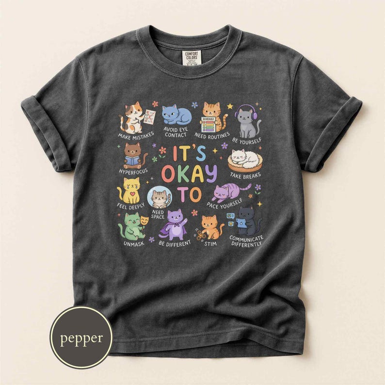 May include: A charcoal gray t-shirt featuring a central graphic of various colorful cartoon cats with the text IT'S OKAY TO, accompanied by phrases like MAKE MISTAKES, AVOID EYE CONTACT, NEED ROUTINES, BE YOURSELF, HYPERFOCUS, NEED SPACE, TAKE BREAKS, UNMASK, BE DIFFERENT, STIM, and COMMUNICATE DIFFERENTLY.