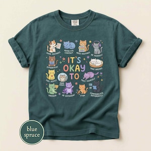 May include: A dark teal short-sleeve t-shirt featuring a graphic of various cartoon cats with the text "IT'S OKAY TO" surrounded by phrases like "AVOID EYE CONTACT," "BE YOURSELF," "HYPERFOCUS," "FEEL DEEPLY," "NEED SPACE," "PACE," "STIM," and "COMMUNICATE DIFFERENTLY."